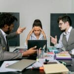 Dwóch mężczyzn kłóci się w biurze, kobieta sfrustrowana patrzy w laptop