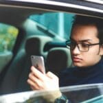 Młody mężczyzna w okularach korzysta ze smartfona w zaparkowanym aucie